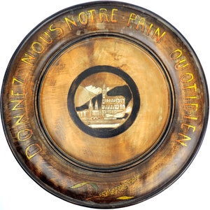 Op de afbeelding: Een ronde houten plaat met een donkerbruine afwerking en een verhoogde rand. De plaat heeft een geschilderd beeld van een gebouw met een zwarte cirkel eromheen. De woorden "DONNEZ NOUS NOTRE PAIN QUOTIDIEN" zijn rond de rand geschilderd in geel en oranje.