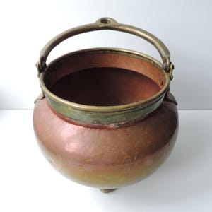 May include: A copper cauldron with a brass handle and rim. The pot has a rounded body and three small feet. The copper has a warm, reddish-brown hue, with a brass rim and handle. The cauldron is likely used for cooking or decorative purposes.
