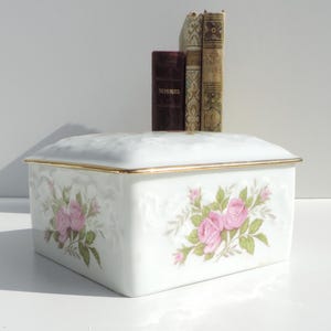 Pu&ograve; includere: Scatola in porcellana bianca con un bordo dorato e un motivo floreale con rose rosa. La scatola ha un coperchio con cerniera.