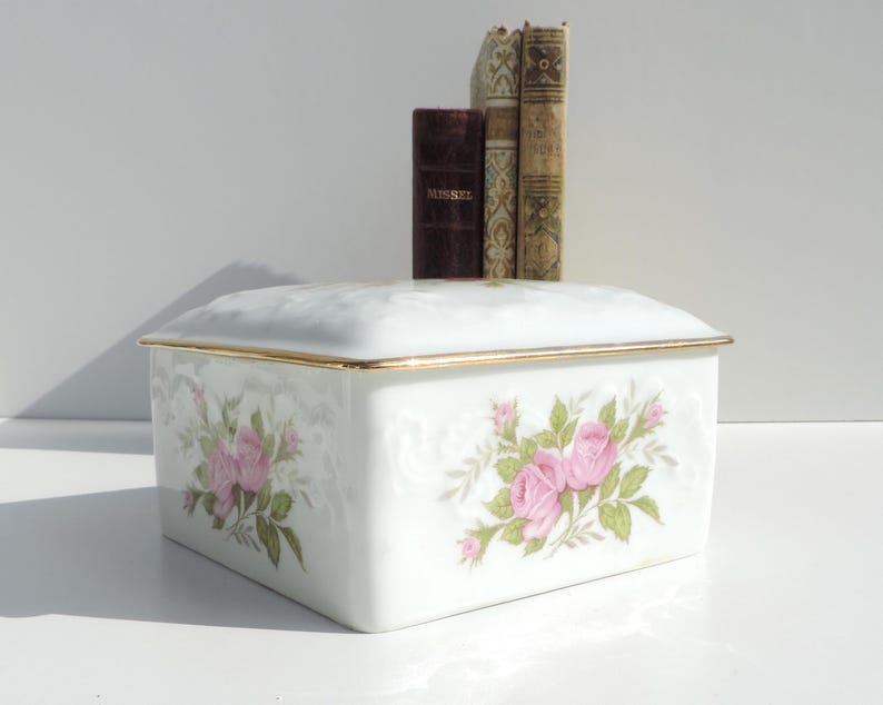 Pu&ograve; includere: Scatola in porcellana bianca con un bordo dorato e un motivo floreale con rose rosa. La scatola ha un coperchio con cerniera.