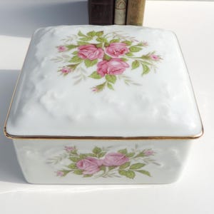 Pu&ograve; includere: Scatola portaoggetti in porcellana bianca con bordo dorato e un motivo floreale con rose rosa e foglie verdi.