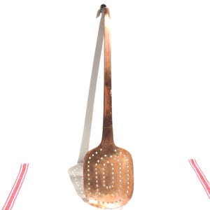 May include: A long-handled copper skimmer with a flat, rectangular head featuring rounded corners and a grid of small, circular perforations. The metal surface displays a warm, metallic copper tone with a slight patina and a curved hook at the top.