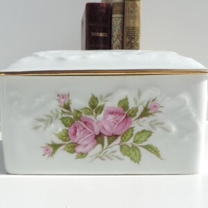 Pu&ograve; includere: Scatola in porcellana bianca con un bordo dorato e un motivo floreale con rose rosa. La scatola ha un coperchio rialzato e testurizzato.