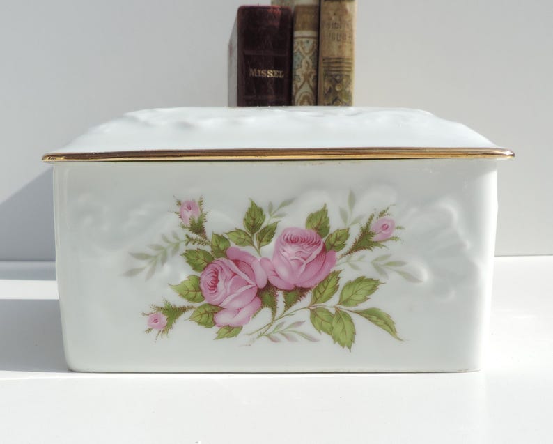 Pu&ograve; includere: Scatola in porcellana bianca con un bordo dorato e un motivo floreale con rose rosa. La scatola ha un coperchio rialzato e testurizzato.