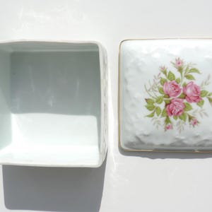 Pu&ograve; includere: Scatola quadrata in porcellana bianca con coperchio. Il coperchio presenta un motivo floreale con rose rosa e foglie verdi. La scatola &egrave; vuota.
