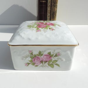 Pu&ograve; includere: Scatola portaoggetti in porcellana bianca con bordo dorato e un motivo floreale con rose rosa. Il coperchio ha un design in rilievo.