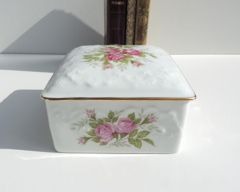 Pu&ograve; includere: Scatola portaoggetti in porcellana bianca con bordo dorato e un motivo floreale con rose rosa. Il coperchio ha un design in rilievo.