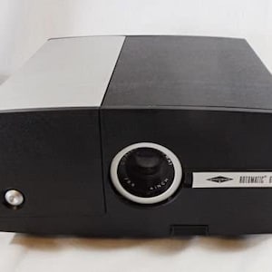 May include: A vintage black and silver slide projector with a prominent lens and the text "4 INCH". The projector has a rectangular shape with a silver top panel and the text "ROTOMATIC 660".