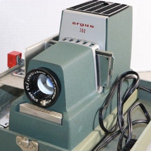 May include: A green Argus 300 slide projector with a black cord and a carrying case. The projector has a lens with a black rim and the text "Argus f:3.3" printed on it.