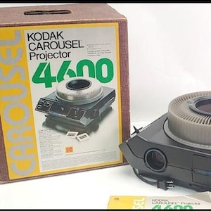 May include: A Kodak Carousel 4600 projector in its original box. The projector is black with a silver carousel and a lens on the front. The box is brown with a yellow and silver label that says "Kodak Carousel Projector 4600".