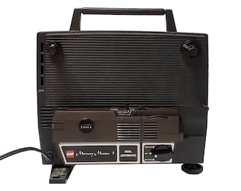 Op de afbeelding: Een zwarte en bruine GAF Memory Master 5 Dual Automatic 8mm-filmprojector met een draagbeugel. De projector heeft een terugspoelknop, een projectieknop en een aan/uit-schakelaar.
