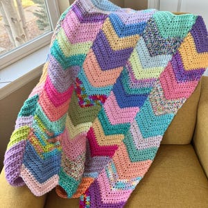 Könnte beinhalten: Eine farbenfrohe, handgefertigte Häkeldecke mit Zickzackmuster. Die Decke weist eine Vielzahl von Pastellfarben auf, darunter Rosa, Lila, Blau und Gelb. Sie ist über einen gelben Stuhl drapiert, mit einem Fenster im Hintergrund.
