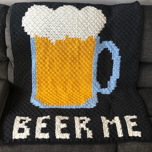 Puede incluir: Una manta de crochet negra con una jarra de cerveza de crochet blanca y amarilla y el texto "BEER ME" en blanco.