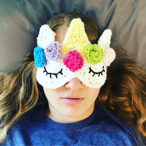 Könnte beinhalten: Eine weiße gehäkelte Schlafmaske in Form eines Einhorns mit bunten Blumen. Die Maske hat schwarze Wimpern und ein weißes Horn.