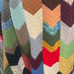 Könnte beinhalten: Eine handgefertigte Decke mit Zickzackmuster in verschiedenen Farben, darunter Blau, Grün, Rot, Braun und Creme. Die Decke ist mit einem strukturierten Stich gehäkelt, was ihr ein gemütliches und einladendes Aussehen verleiht. Die Decke ist über einer dunklen Oberfläche gefaltet.