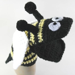 May include: A crocheted bee puppet with black and yellow stripes, white wings, and black eyes. The bee is wearing a white hat.
