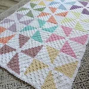 Könnte beinhalten: Eine rechteckige Häkeldecke mit einem sich wiederholenden Windradmuster. Das Design besteht aus weißen Garnquadraten, die sich mit dreieckigen Abschnitten in Pastelltönen wie Gelb, Blau, Lila, Pfirsich, Minzgrün und Rosa abwechseln.