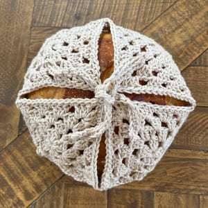 May include: A round loaf of bread wrapped in a beige crocheted bread cover. The cover is tied at the top, and the bread is visible through the openwork design. The bread sits on a wooden surface.
