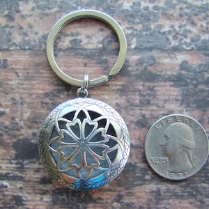 May include: Silver key chain with a round locket featuring a decorative floral design. The locket has a small opening for inserting essential oils or other scents. The key chain is shown next to a US quarter for size comparison.