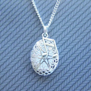 May include: A silver-colored necklace with a detailed, openwork pendant. The pendant is oval-shaped, with a Ferris wheel design inside. The chain is a delicate curb link. The background is a blue and gray patterned surface.