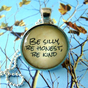 May include: A silver necklace with a round pendant featuring the words "Be silly, be honest, be kind" written in black ink on a cream background.