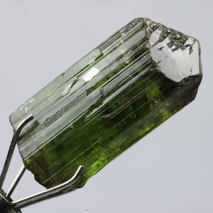 Könnte beinhalten: Ein grüner, facettierter, länglicher Kristall mit Streifen. Der Kristall wird in einer Metallklemme gehalten.