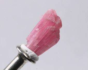 May include: A pink, elongated, crystal specimen mounted on a silver pin. The crystal has a striated, layered appearance.