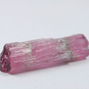 Puede incluir: Un cristal de turmalina rosa y blanco con estrías. El cristal es translúcido y tiene un extremo puntiagudo.