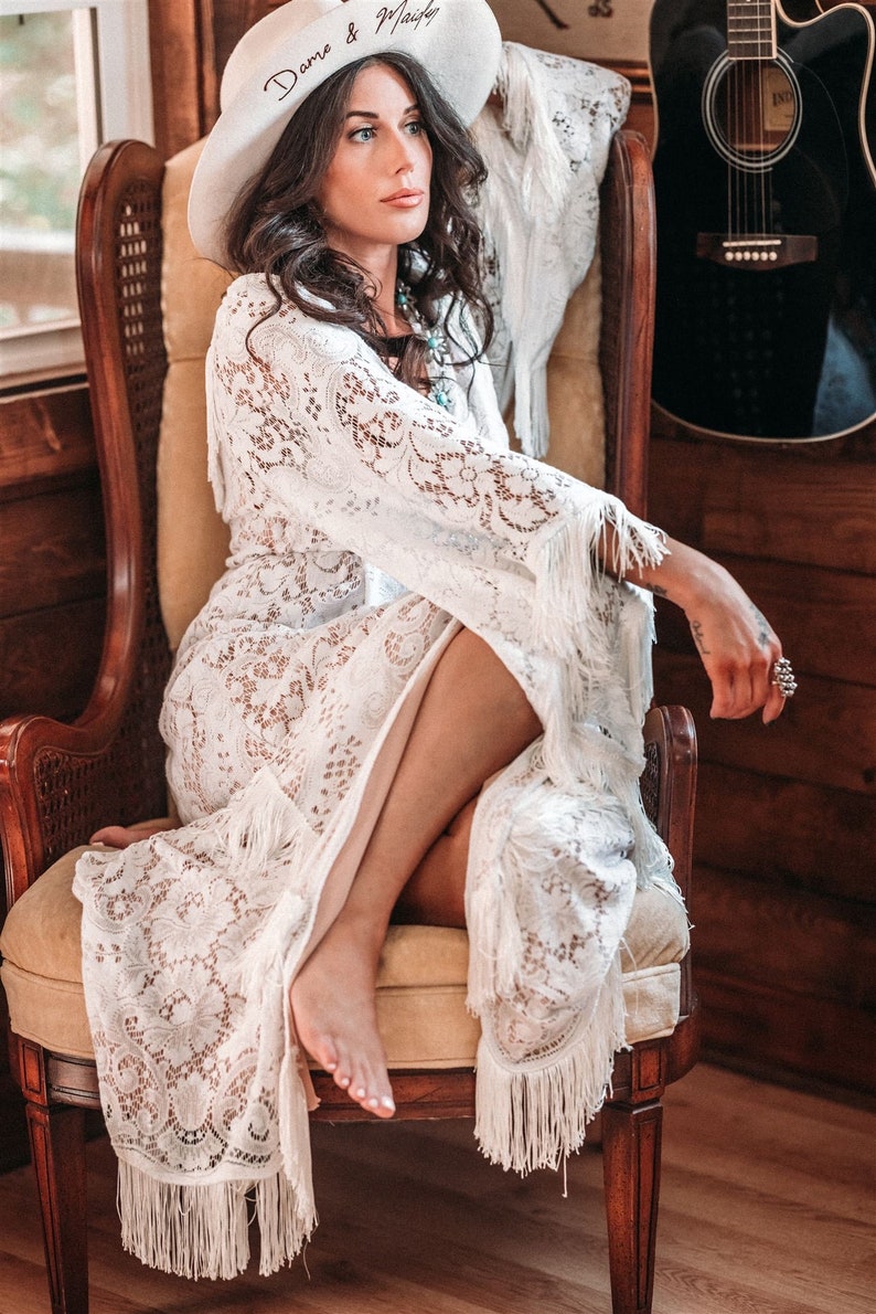 May include: A woman wearing a white lace dress with fringe details sits in a chair. She is wearing a white hat with the text "Domo & Mailey" written on it. The woman is looking to the right of the frame.
