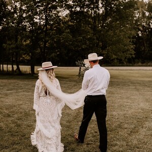 May include: A bride and groom walking away from the camera in a field. The bride is wearing a white lace dress with a long veil and a wide-brimmed hat. The groom is wearing a white shirt, black pants, and a cowboy hat.
