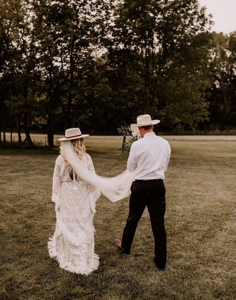 May include: A bride and groom walking away from the camera in a field. The bride is wearing a white lace dress with a long veil and a wide-brimmed hat. The groom is wearing a white shirt, black pants, and a cowboy hat.