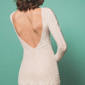 May include: A woman wearing a white lace dress with a low back and a floral crown. The dress has a fitted silhouette and long sleeves. The crown is made of pink and white flowers.