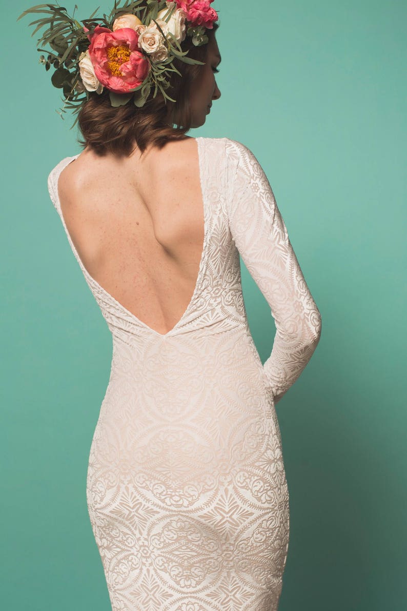 May include: A woman wearing a white lace dress with a low back and a floral crown. The dress has a fitted silhouette and long sleeves. The crown is made of pink and white flowers.