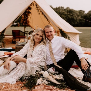 May include: A couple poses for a wedding photo in front of a white tent decorated with greenery. The bride wears a white lace dress and the groom wears a white shirt and black pants with a patterned tie. They are sitting on a colorful rug with a bouquet of flowers in front of them.