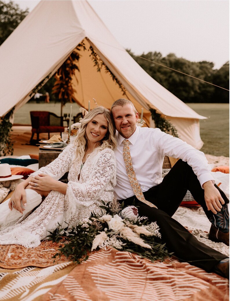 May include: A couple poses for a wedding photo in front of a white tent decorated with greenery. The bride wears a white lace dress and the groom wears a white shirt and black pants with a patterned tie. They are sitting on a colorful rug with a bouquet of flowers in front of them.