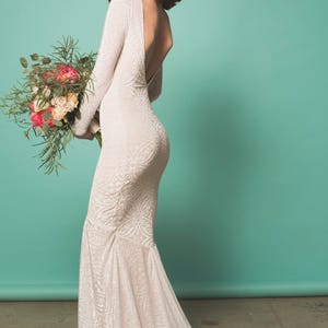 May include: A white lace wedding dress with a low back and long sleeves. The dress has a fitted bodice and a flowing skirt. The model is holding a bouquet of pink and white flowers.