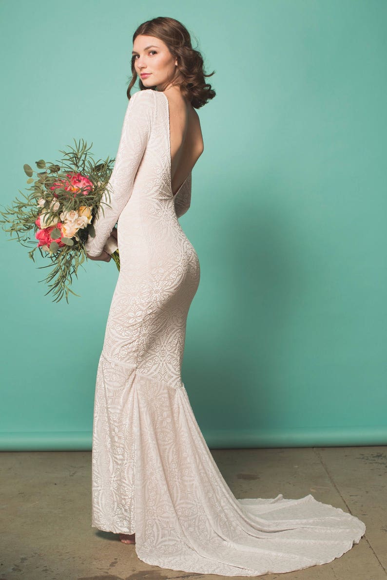 May include: A white lace wedding dress with a low back and long sleeves. The dress has a fitted bodice and a flowing skirt. The model is holding a bouquet of pink and white flowers.