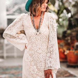 May include: A white lace maxi dress with long sleeves and a V-neckline. The dress features a delicate floral pattern and is paired with a teal blue hat.
