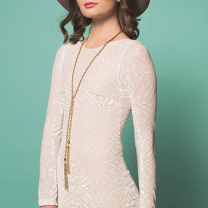 May include: A woman wearing a white lace dress with a gold chain necklace and a brown hat with a pink flower on top. The background is a light turquoise color.