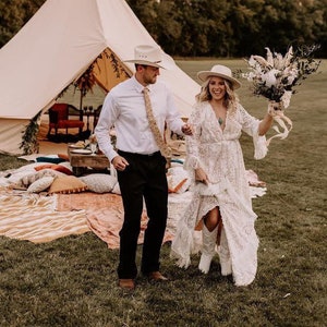 May include: A bride and groom stand in a field with a white tent and a bouquet of dried flowers. The bride is wearing a white lace dress and a white hat. The groom is wearing a white shirt, a brown tie, and a brown hat.
