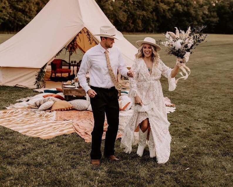 May include: A bride and groom stand in a field with a white tent and a bouquet of dried flowers. The bride is wearing a white lace dress and a white hat. The groom is wearing a white shirt, a brown tie, and a brown hat.