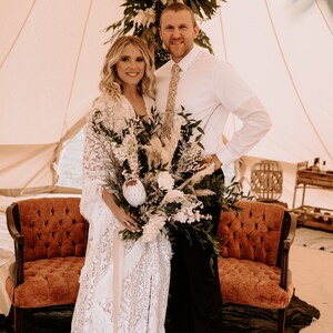 May include: A bride and groom pose for a wedding photo in a white tent decorated with greenery. The bride is wearing a white lace dress and holding a bouquet of white and green flowers. The groom is wearing a white shirt and a tie.