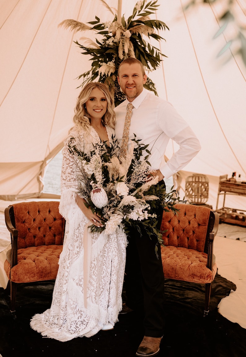 May include: A bride and groom pose for a wedding photo in a white tent decorated with greenery. The bride is wearing a white lace dress and holding a bouquet of white and green flowers. The groom is wearing a white shirt and a tie.