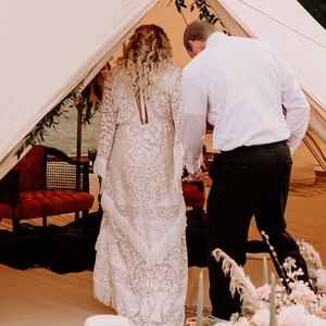May include: A couple in a white tent, the bride is wearing a white lace dress with a long train, the groom is wearing a white shirt and black pants.