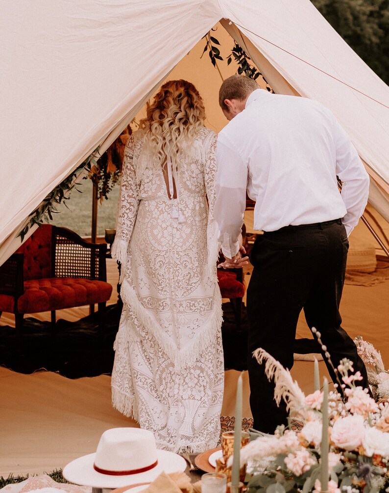 May include: A couple in a white tent, the bride is wearing a white lace dress with a long train, the groom is wearing a white shirt and black pants.