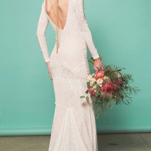 May include: A white lace wedding dress with a long train and a deep V-neck back. The dress has a fitted bodice and a flowing skirt. The model is holding a bouquet of pink and white flowers.