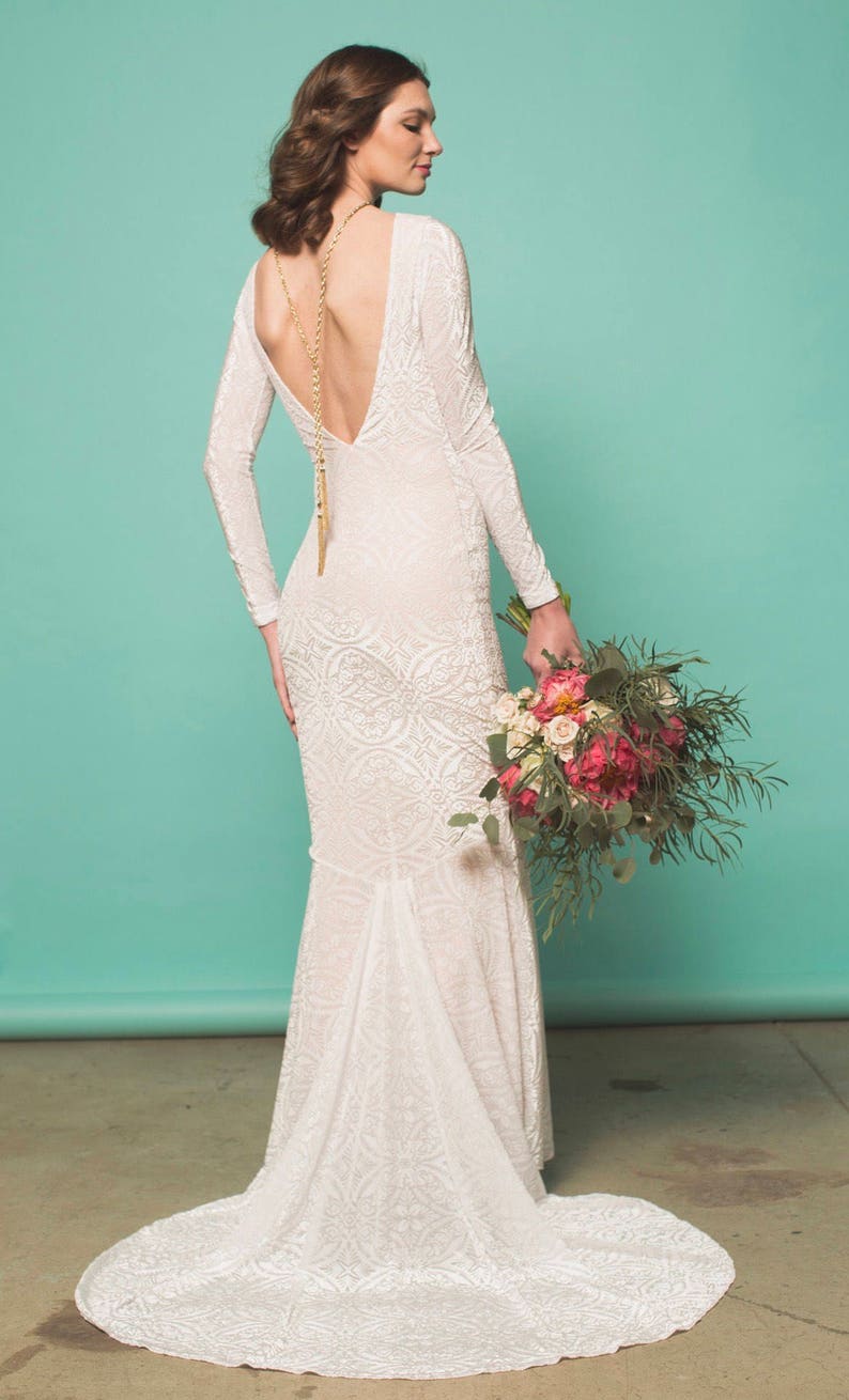 May include: A white lace wedding dress with a long train and a deep V-neck back. The dress has a fitted bodice and a flowing skirt. The model is holding a bouquet of pink and white flowers.