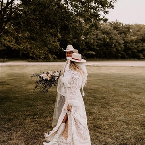 May include: A bride and groom in white cowboy hats and white clothing stand in a grassy field. The bride is wearing a white lace dress with a long veil and white cowboy boots. The groom is wearing a white shirt and white pants. They are both smiling and looking at each other.