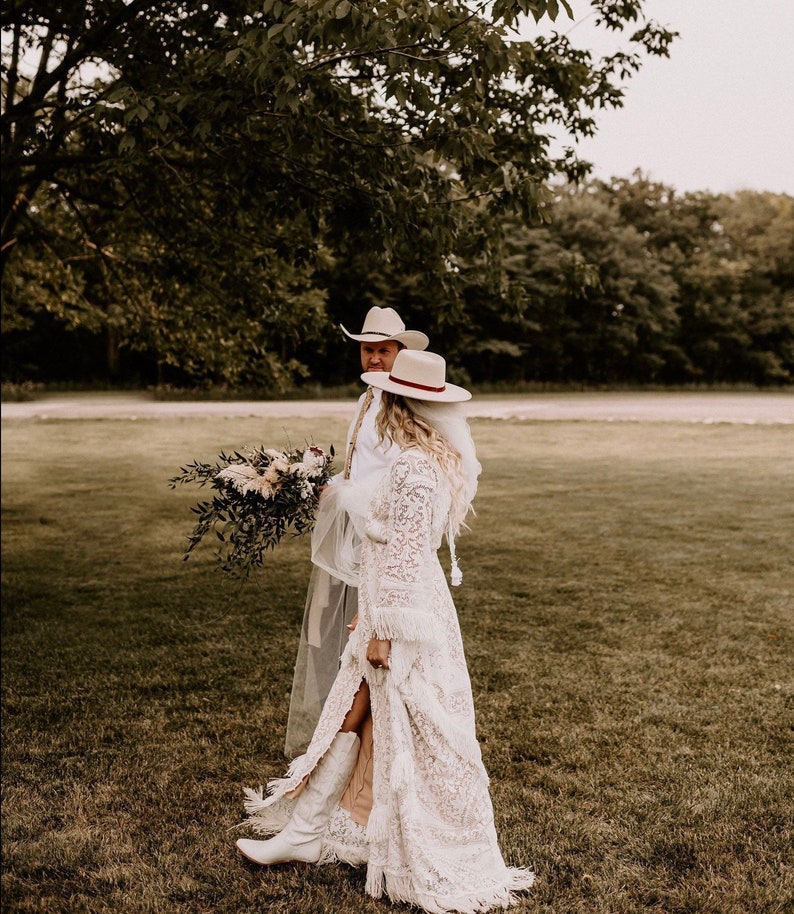 May include: A bride and groom in white cowboy hats and white clothing stand in a grassy field. The bride is wearing a white lace dress with a long veil and white cowboy boots. The groom is wearing a white shirt and white pants. They are both smiling and looking at each other.