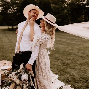 May include: A couple in a field, wearing white cowboy hats. The woman is wearing a white lace dress and a veil. The man is wearing a white shirt and black pants. They are holding a bouquet of flowers.